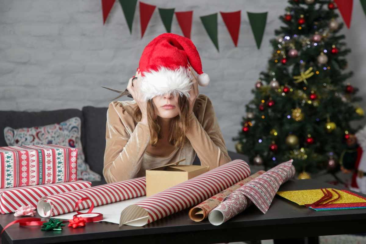 Weihnachtsstress: Frau sitzt mit Weihnachtsmütze bis über die Augen heruntergezogen an einem Tisch voller Geschenkpapier
